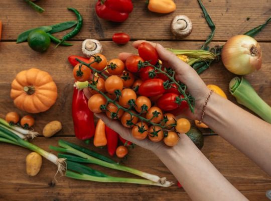 Hands holding cherry tomatoes surrounded by fresh vegetables on a rustic wooden surface.