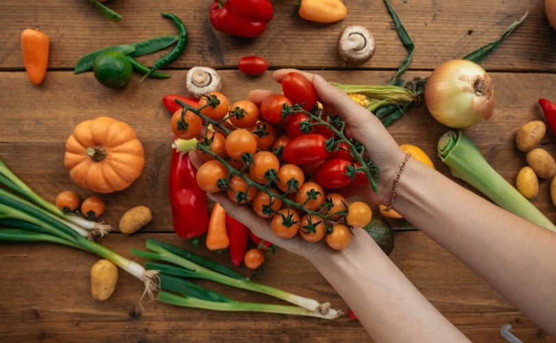 Hands holding cherry tomatoes surrounded by fresh vegetables on a rustic wooden surface.
