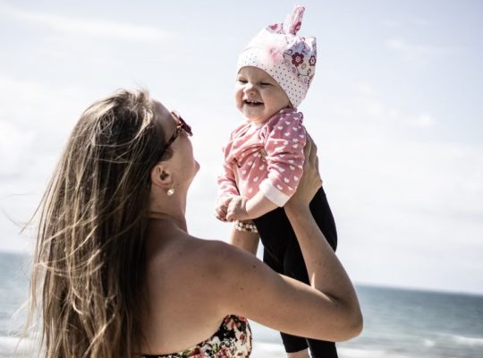 A happy mother lifting her smiling baby against a scenic beach backdrop.