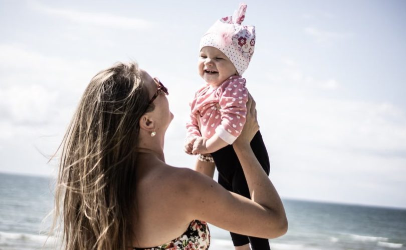 A happy mother lifting her smiling baby against a scenic beach backdrop.